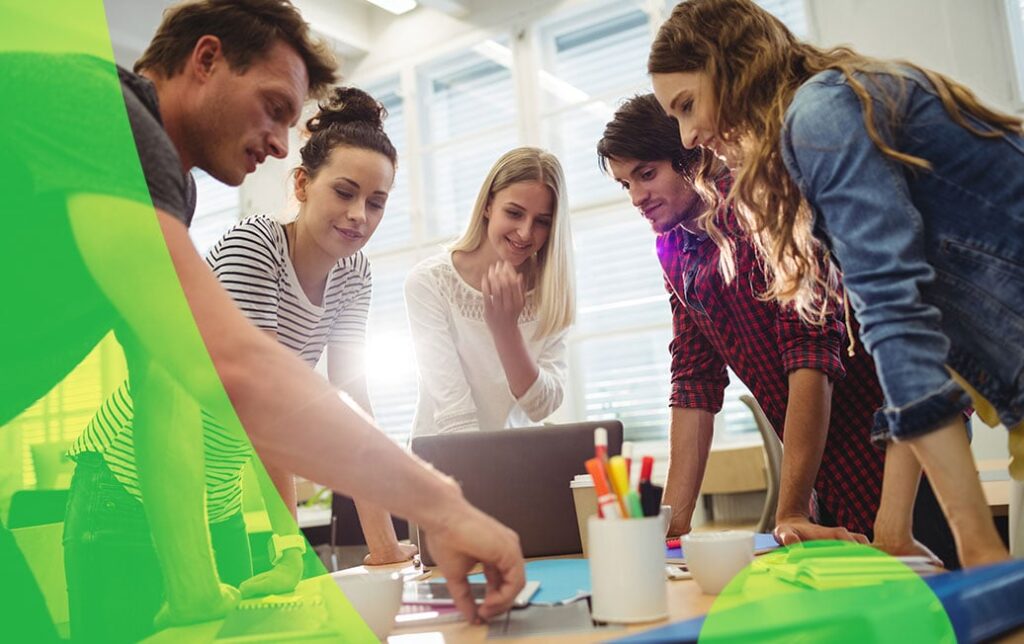 Zdjęcie przedstawia grupę młodych ludzi pochylonych nad stołem. Na stole znajduje się laptop oraz artykuły papiernicze. Osoby na zdjęciu są uśmiechnięte, niektóre z nich patrzą na siebie lub wpatrują się w przedmioty na stole.